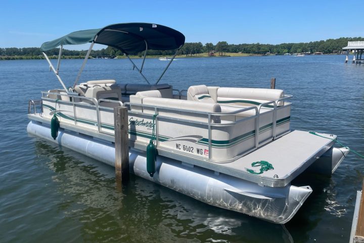 Pontoon boat with canopy docked on a lake with clear sky.