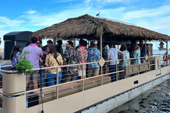 Group of people in tropical shirts on a large boat with thatched roof, under a blue sky.