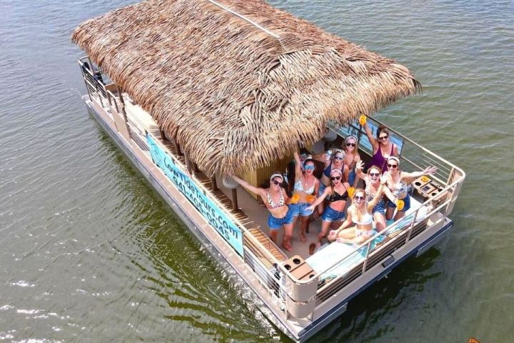 Group of people on a pontoon boat with a thatched roof, raising glasses and smiling on water.