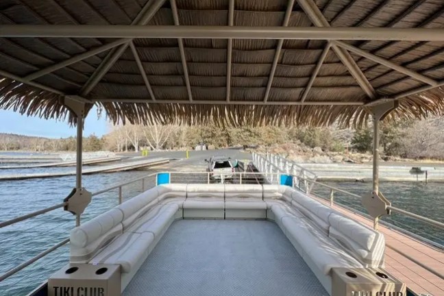 Empty pontoon boat with tiki roof docked by a lake.