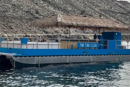 Blue pontoon boat with a thatched roof named 'The Tiki Cloud' on a lake near a rocky shore.