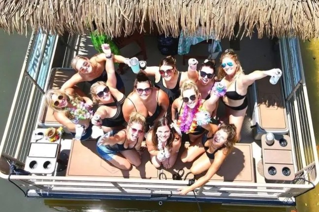Group of women in swimsuits on a boat, holding drinks and wearing sunglasses.